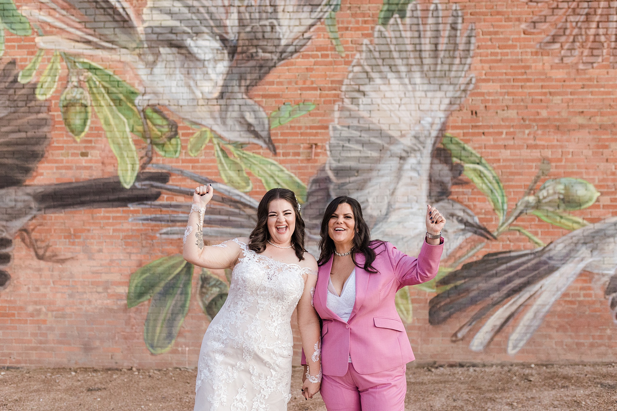 Two Caucasian brides holding hands, raising their opposite hands in celebration, and cheering in front of an outdoor painted mural at Artspace 111 in Fort Worth, Texas. One bride is wearing a detailed, floral, white wedding dress, a floral hairpiece, and a pearl necklace. The other bride is wearing a pink suit, white dress shirt, jeweled necklace, and a jeweled watch. The mural is of birds and flowers painted onto red brick.