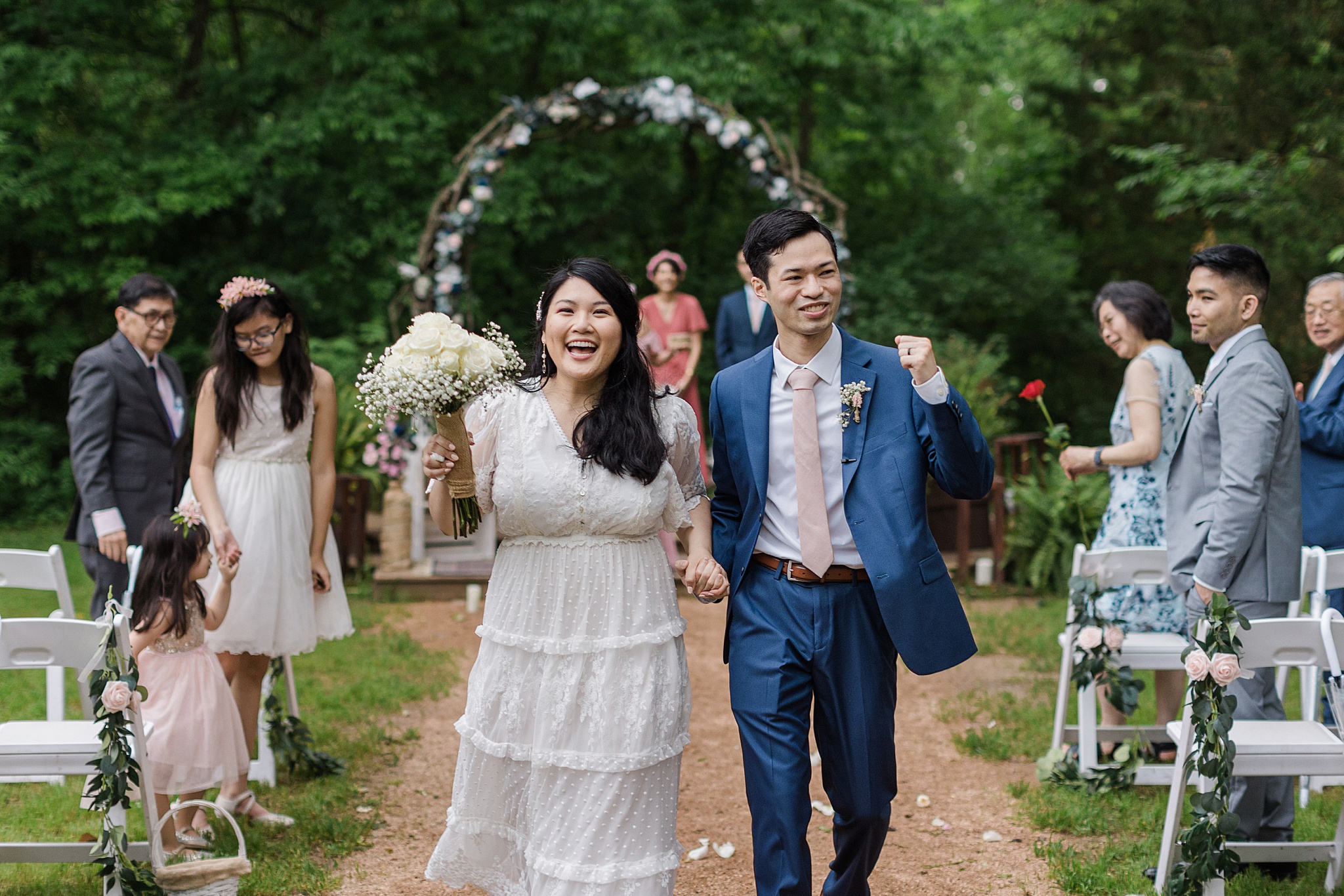 Asian bride and groom walking down the aisle after the conclusion of their wedding ceremony. They are both smiling and celebrating while holding hands and reaching up into the air. The bride is wearing a detailed white dress, a floral hair piece, and holds a bouquet of white roses. The groom is wearing a blue suit, white dress shirt, pale pink tie, and boutonniere. They are surrounded by wedding guests who are also celebrating. In the background is the wedding officiant and large wooden wedding arch covered in flowers in front of a wooded background.