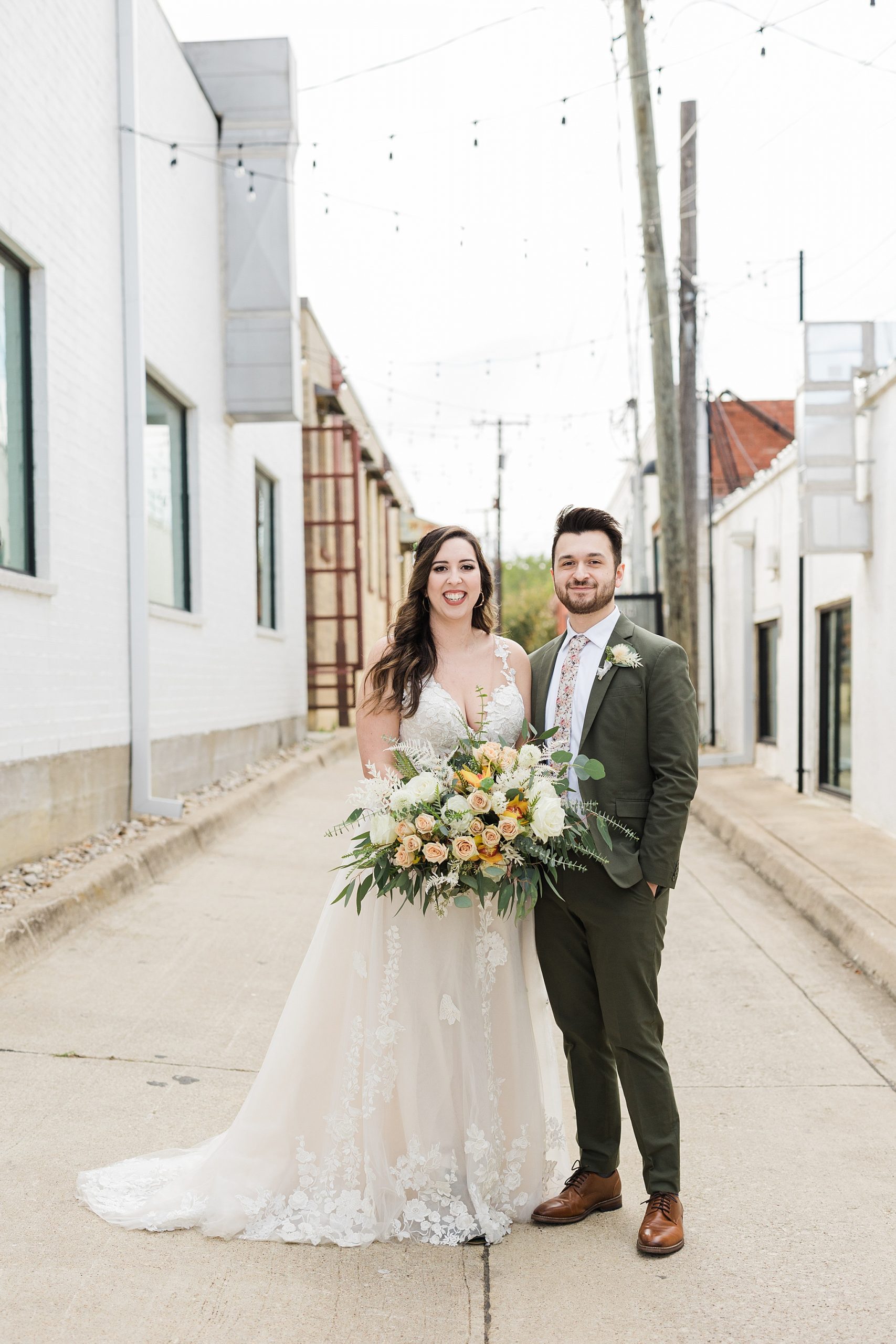 A bride and groom smiling and posing happily for a portrait before their wedding at The 4 Eleven in Fort Worth, Texas. The bride is wearing a decorative, floral, white wedding dress, green shoes, a floral headpiece, and is a holding a large bouquet of flowers. The groom is wearing a grey suit, white dress shirt, decorative floral tie, a boutonniere, and a watch.
