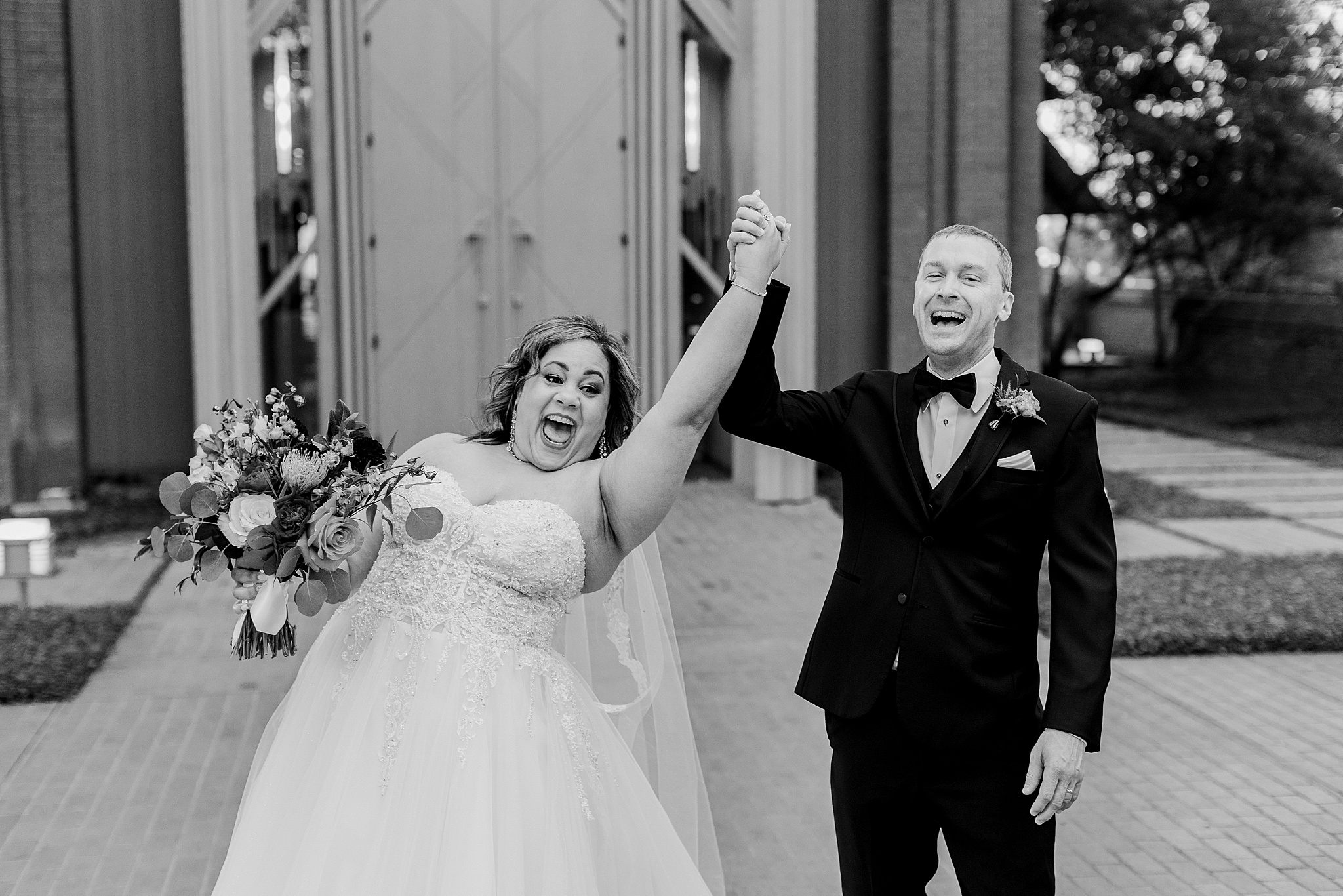 A black and white candid photo of an African American bride and a Caucasian groom laughing and holding their holds together and up in the air in front of the Marty Leonard Community Chapel in Fort Worth, Texas. The bride is wearing a flowing, white, highly decorative, wedding dress, a necklace, a bracelet, a long, flowing veil, and is holding a large bouquet. The groom is wearing a black tuxedo with a white pocket square, and an orange and blue boutonniere.